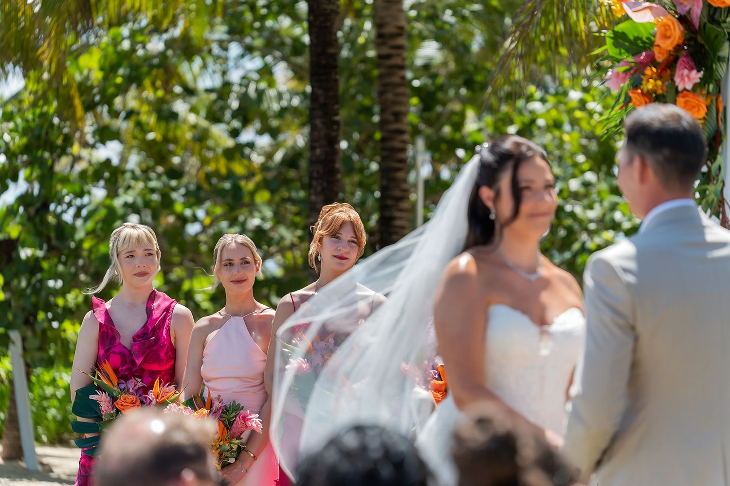 Bridesmaids in pink dresses holding colorful bouquets watch a bride during an outdoor tropical wedding ceremony.