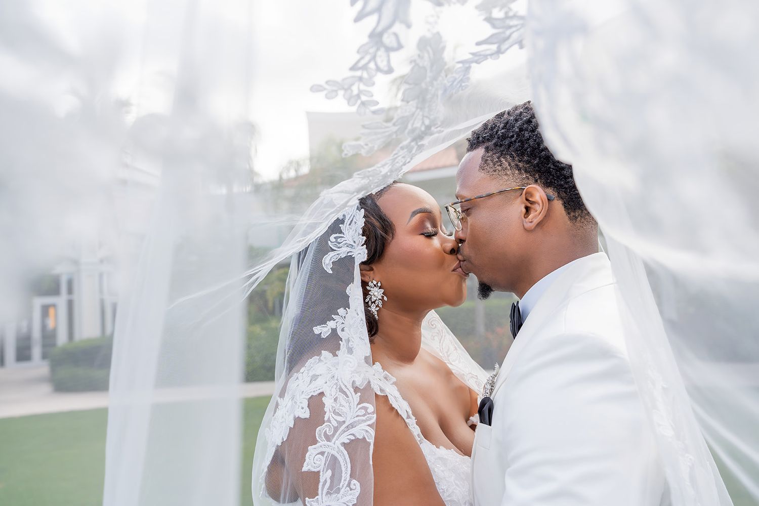 Bride and groom sharing a romantic kiss under a flowing lace wedding veil outdoors.