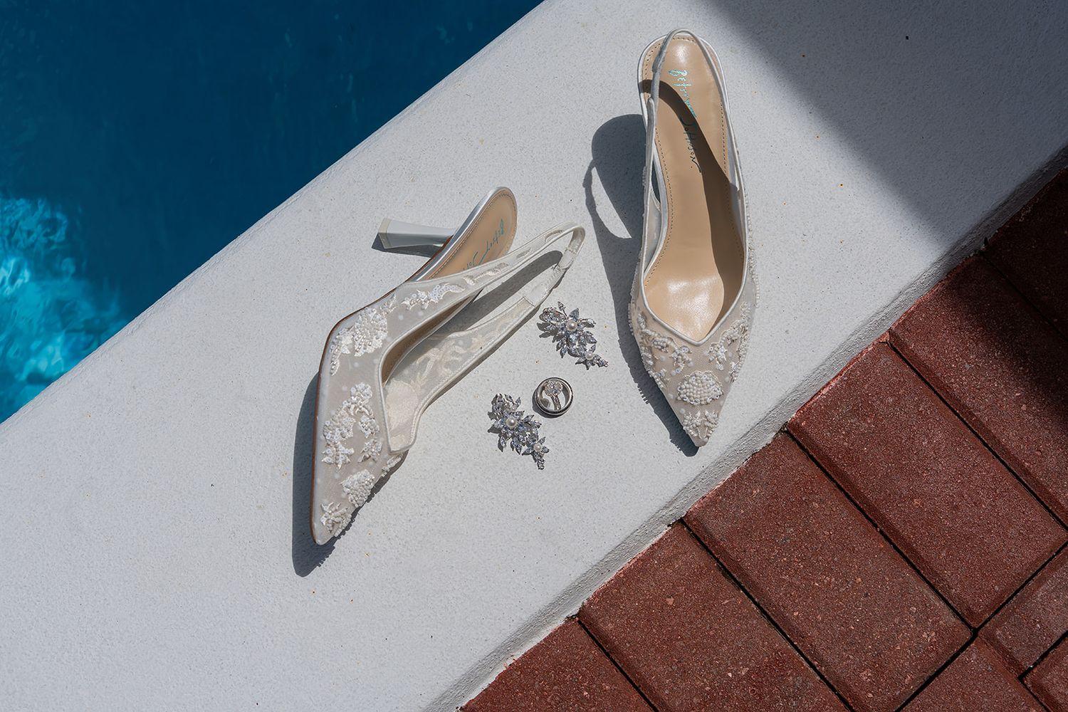 Elegant lace bridal heels and jewelry displayed on white surface near blue pool in natural sunlight.