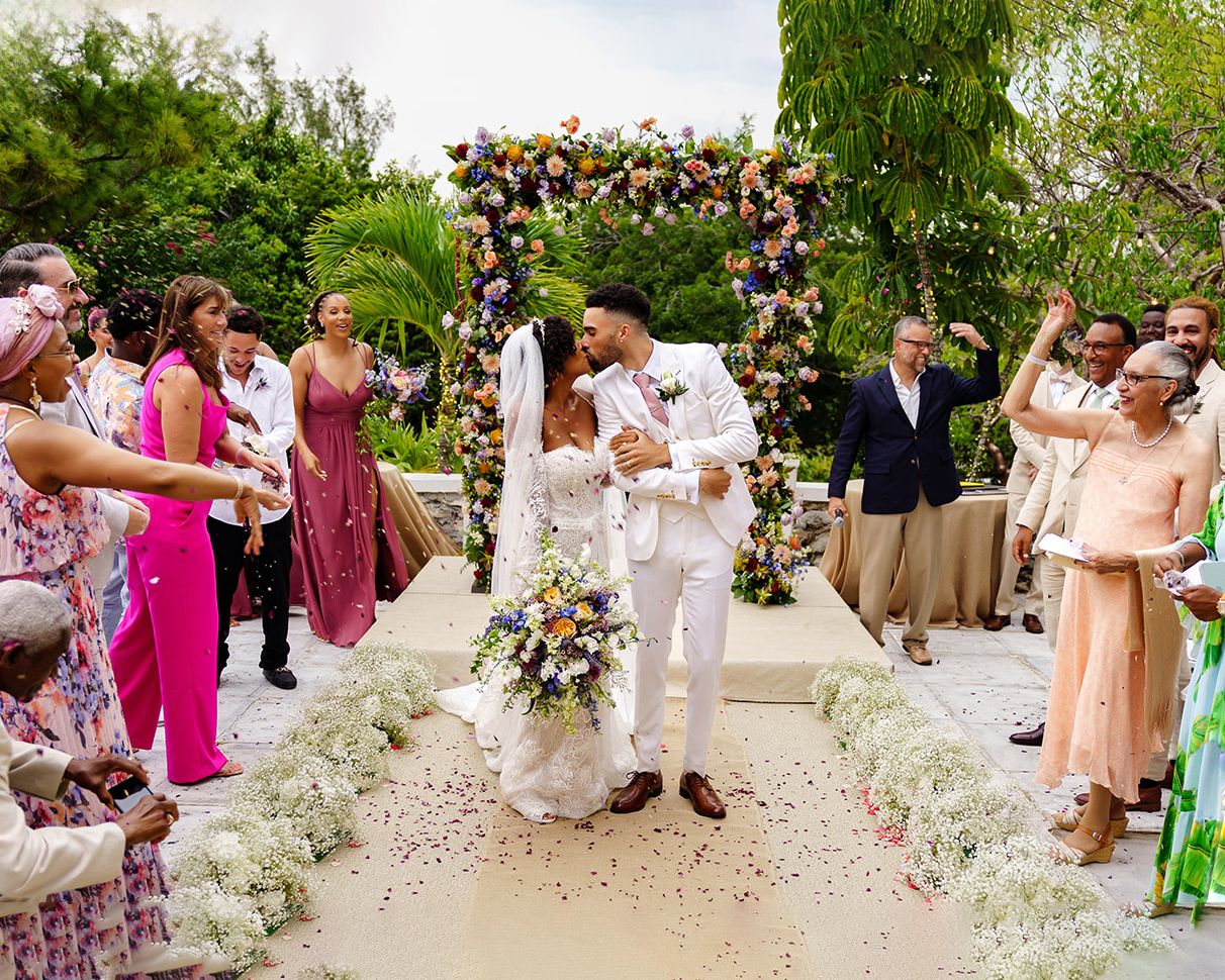 Joyful wedding ceremony with couple kissing under floral arch, guests celebrating with confetti on elegant outdoor aisle.