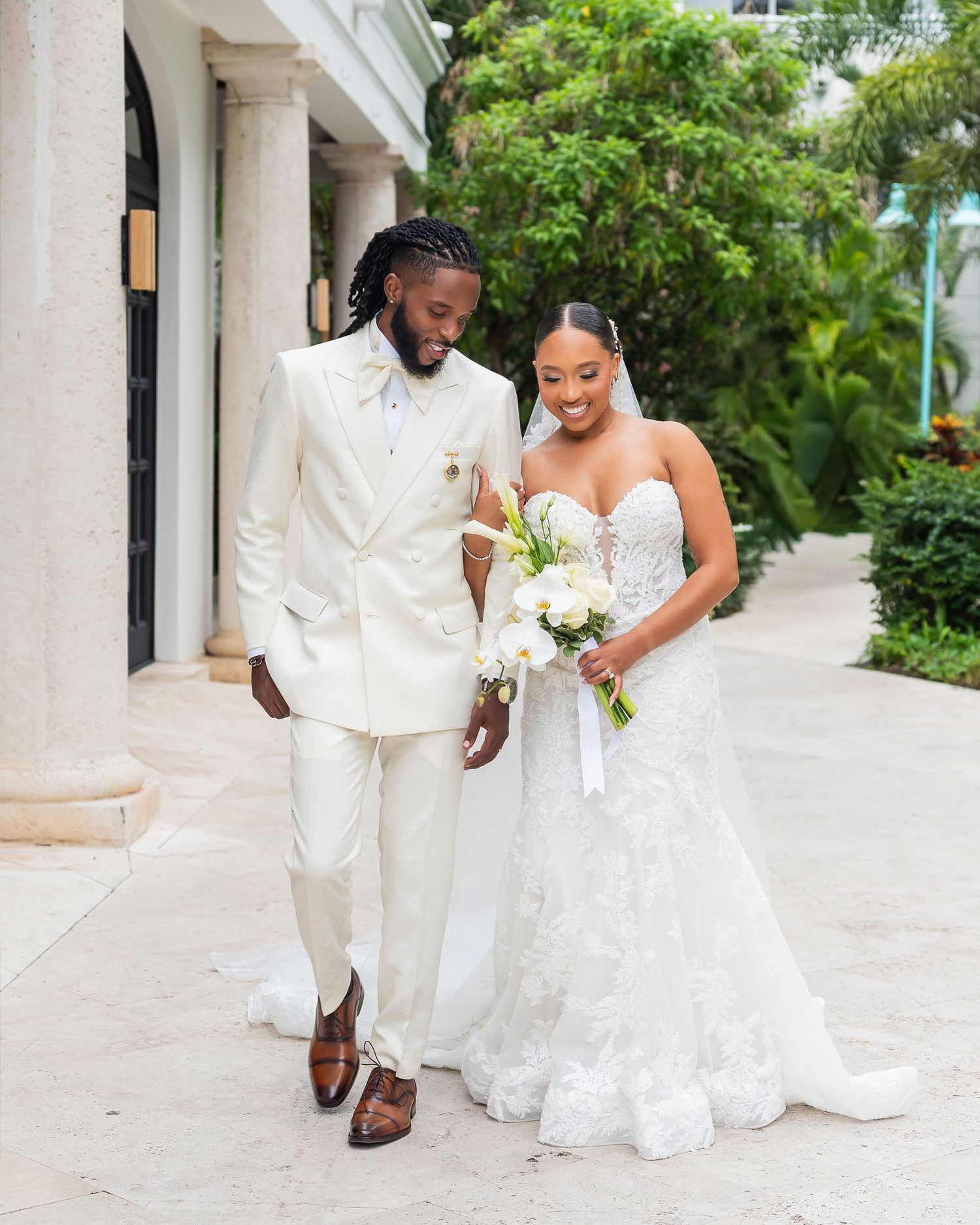 A couple in wedding attire walks together, groom in cream suit, bride in lace gown holding white bouquet.