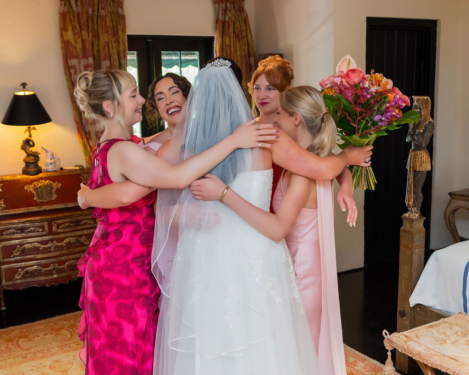 Bride in white gown and veil shares joyful group hug with bridesmaids in pink dresses holding bouquet.