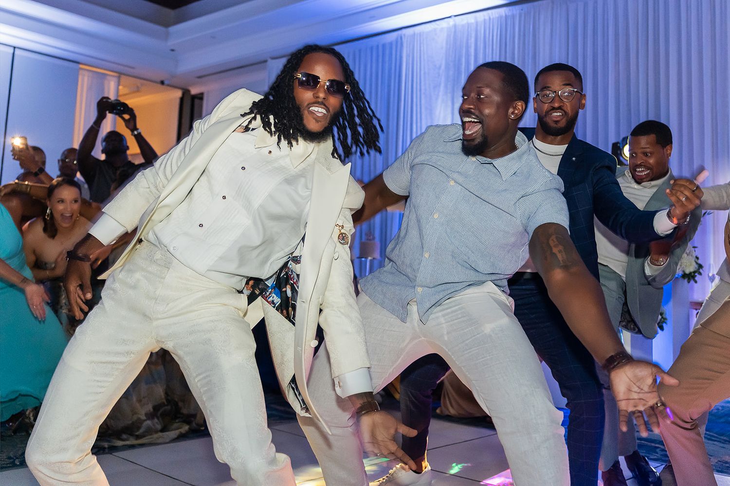 Two men in white suits dance energetically at a formal celebration with guests cheering in the background.