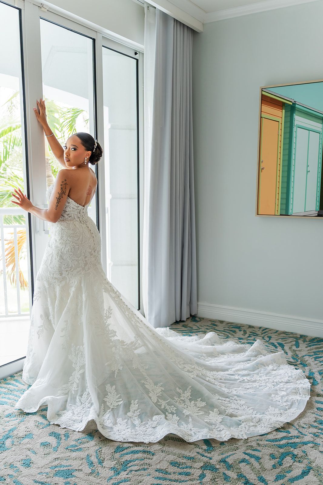 Bride in elegant lace mermaid wedding gown with long train standing by window in tropical hotel room.