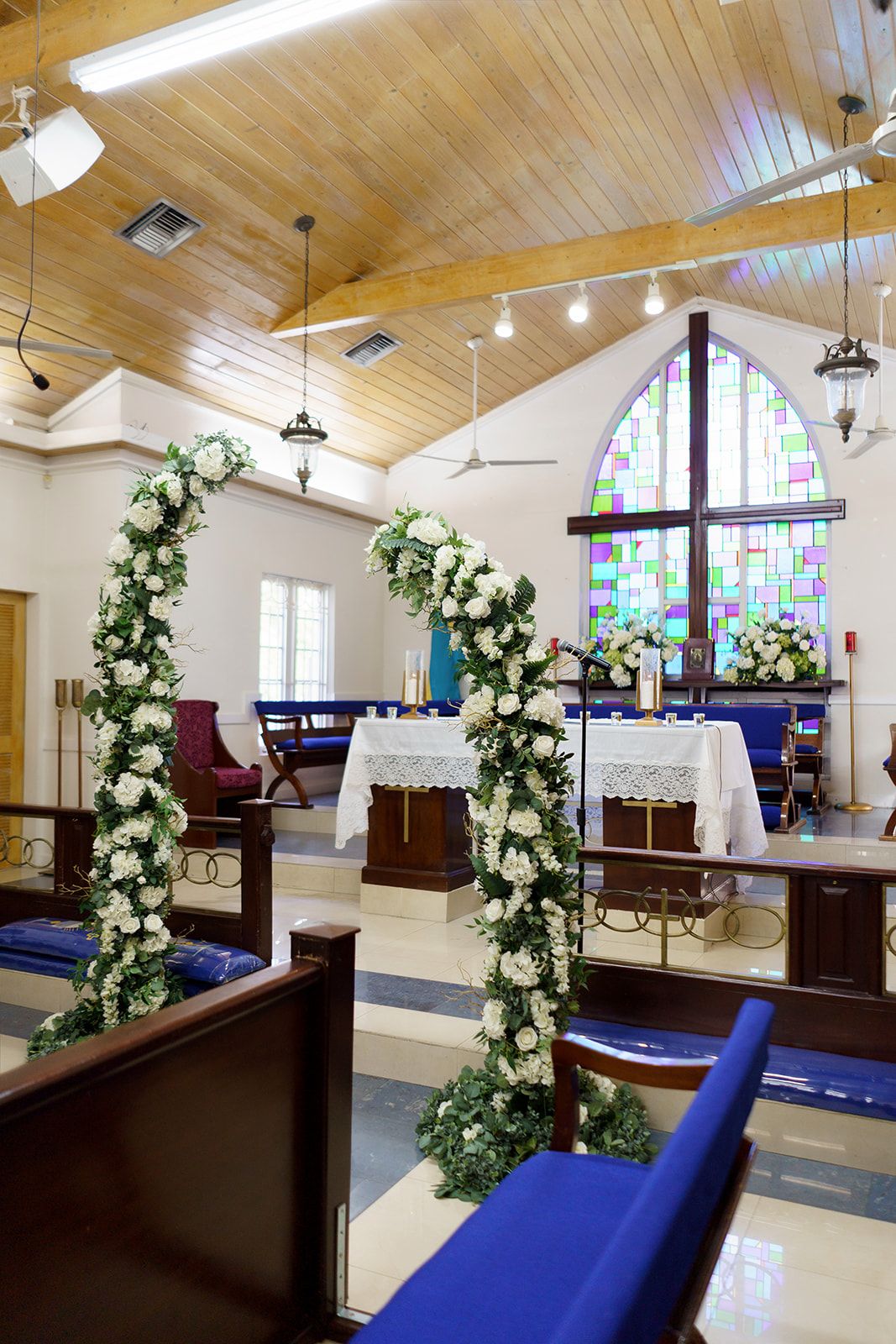 Church interior decorated with white floral arches and greenery for a wedding ceremony, featuring stained glass windows and blue pews.