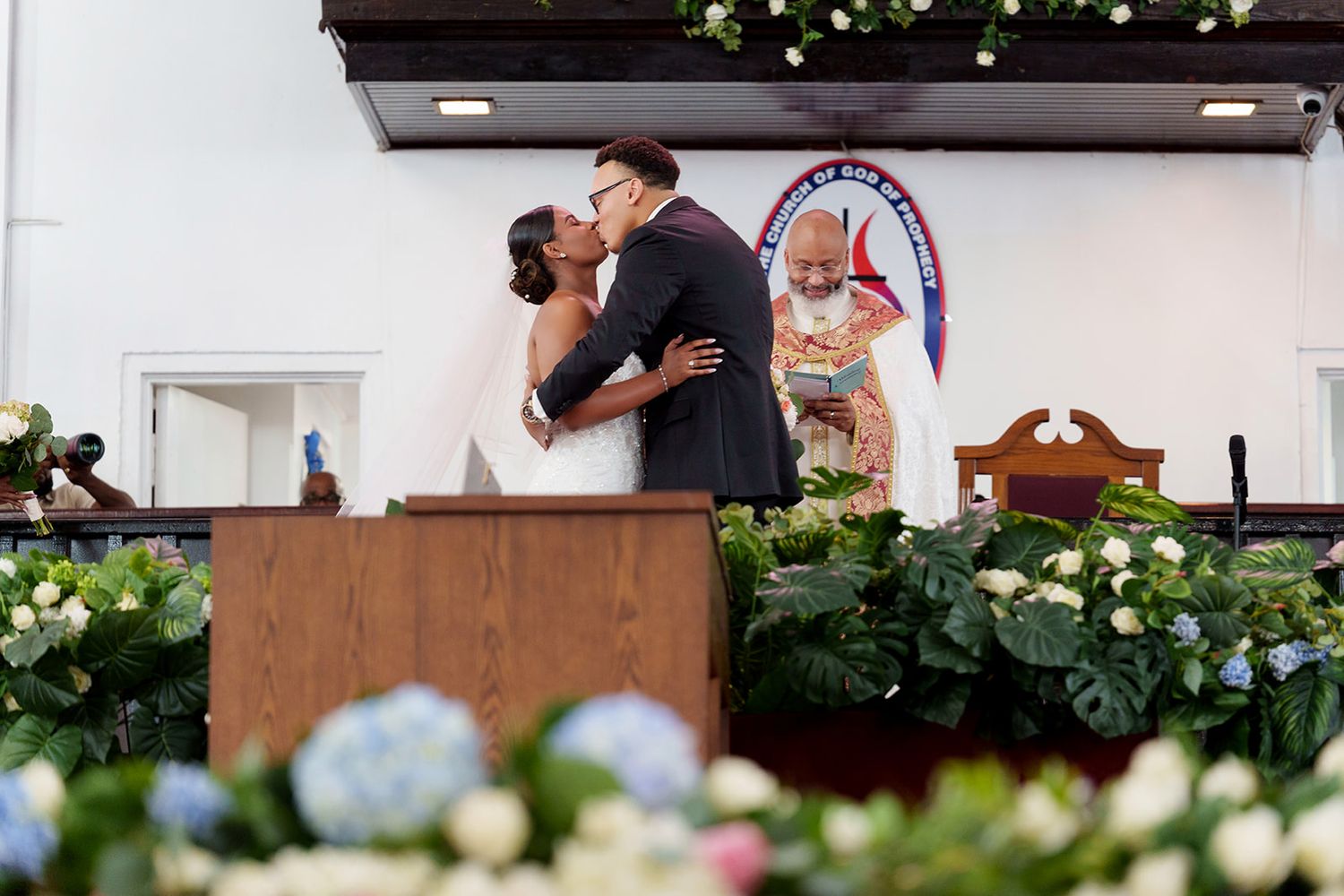 Bride and groom share first kiss at altar surrounded by floral arrangements in church.