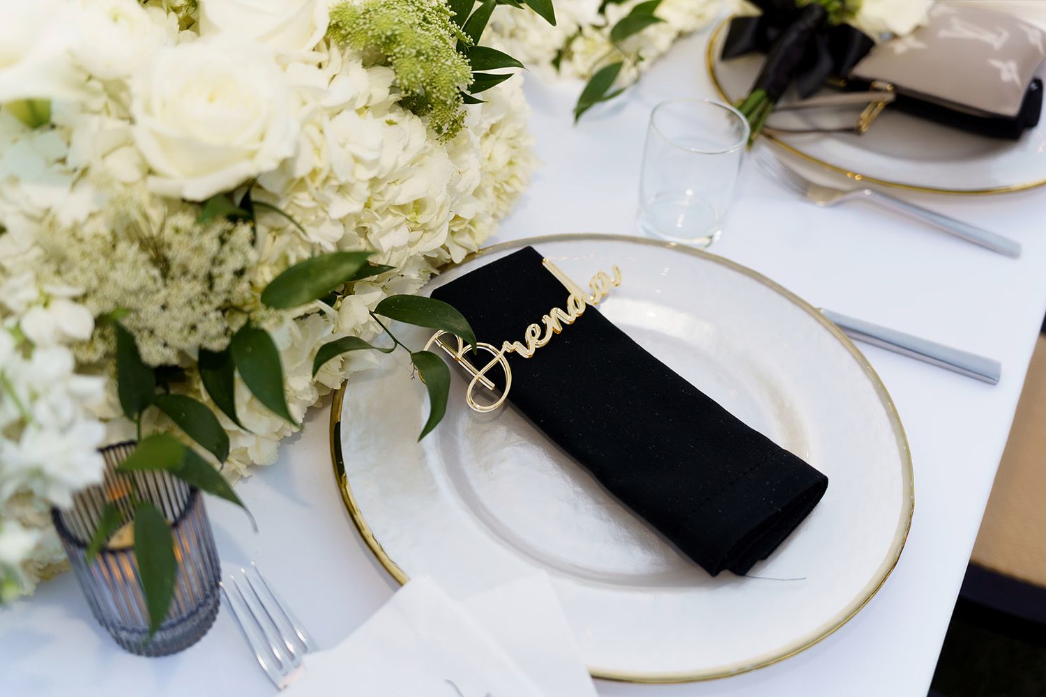 Elegant wedding place setting with gold-rimmed charger, black napkin, cursive name card, and white floral centerpiece.