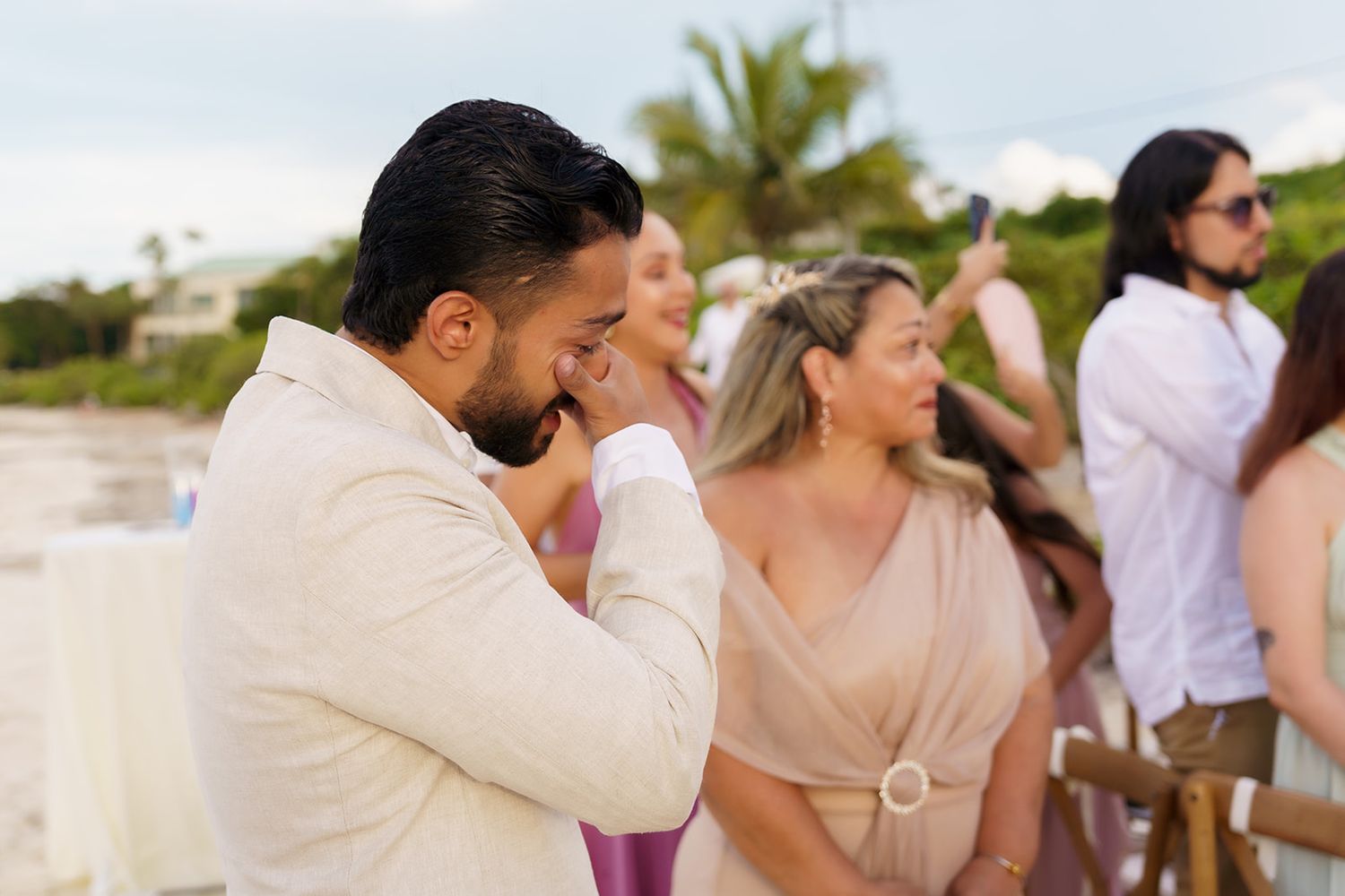 Emotional groom wiping tears at outdoor beach wedding ceremony with bridesmaids and guests in background.