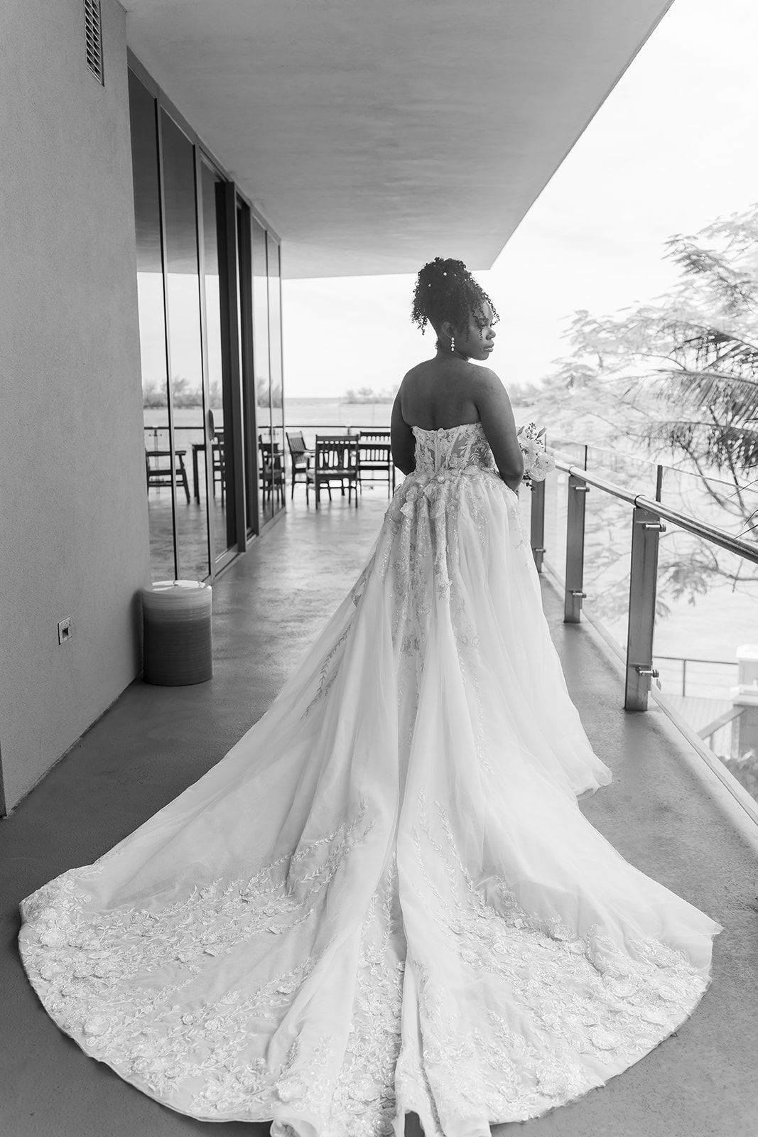 Black and white photo of a bride in an elegant lace wedding gown with a long train on a modern balcony.