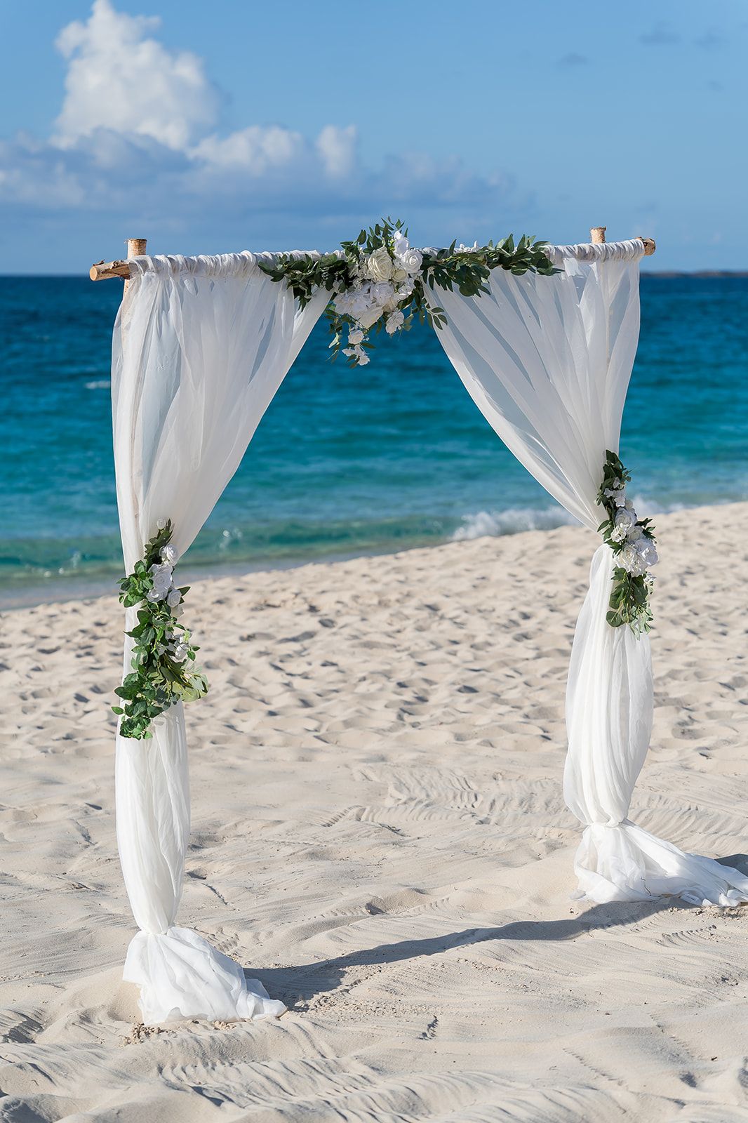 White draped wedding arch decorated with greenery on a sandy beach with turquoise ocean backdrop.