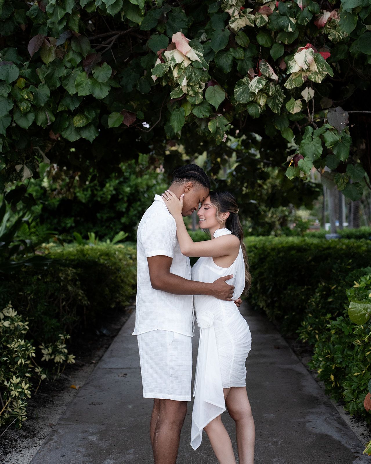 Romantic couple in white outfits embracing on a garden pathway surrounded by lush green foliage.