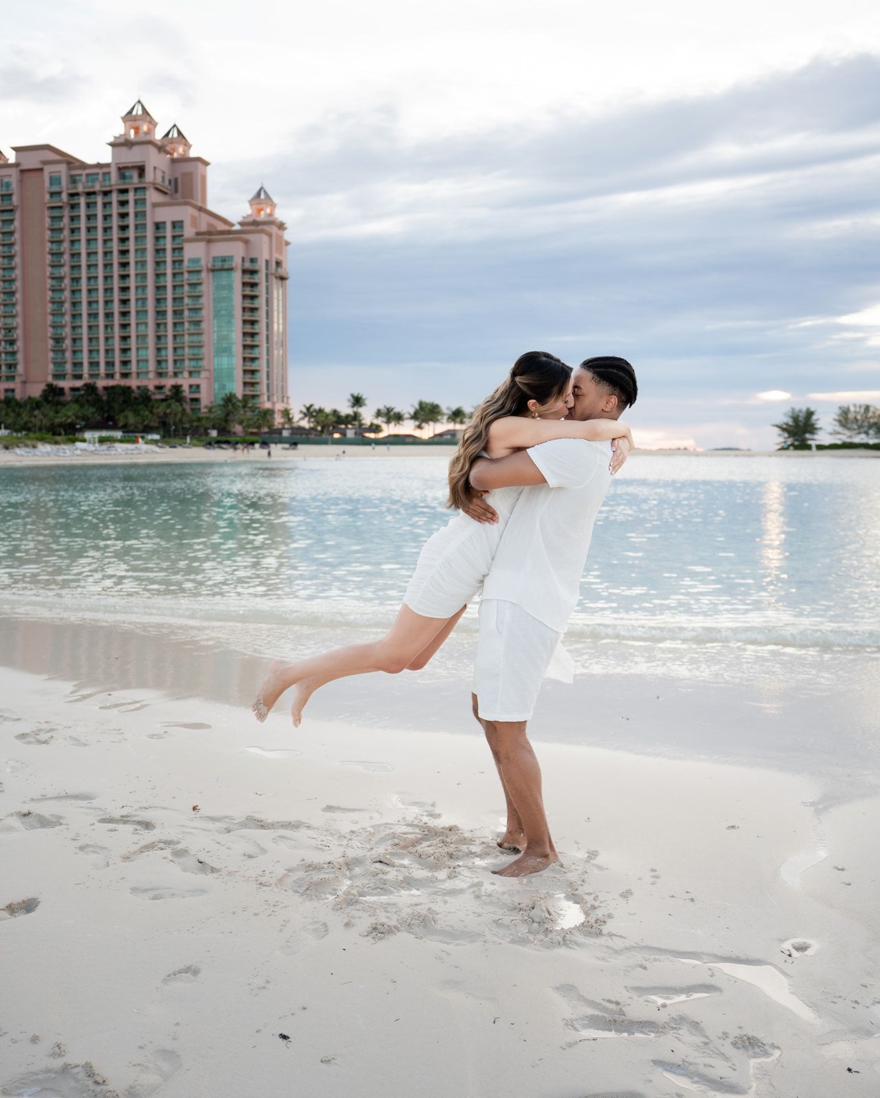 Couple in white outfits embracing on a tropical beach with a pink resort hotel in the background.