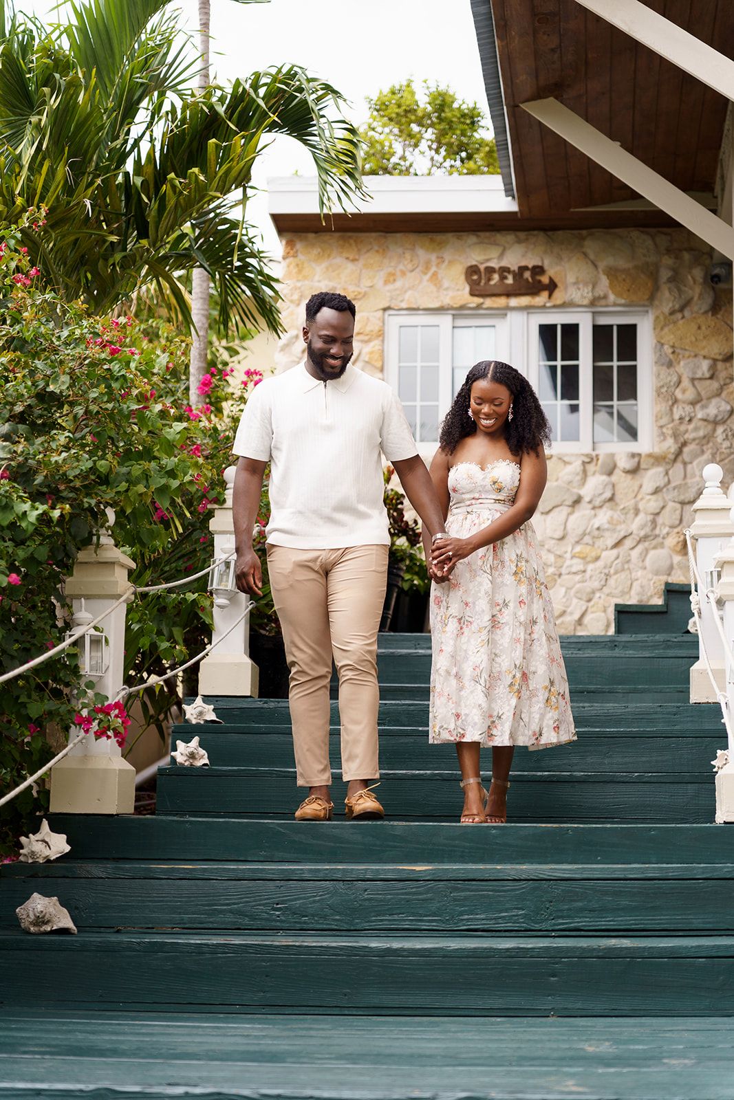 A smiling couple holds hands while walking down green painted outdoor stairs at a tropical resort with lush greenery.
