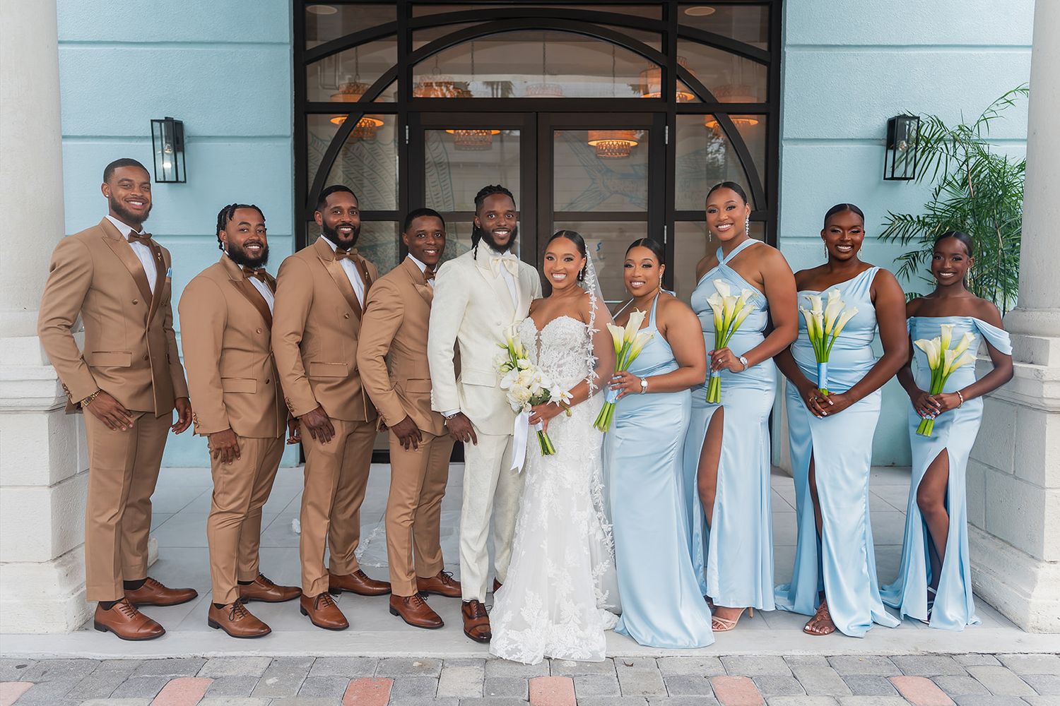 Wedding party poses outdoors, groomsmen in tan suits, bridesmaids in light blue gowns, bride in white lace, groom in white suit.