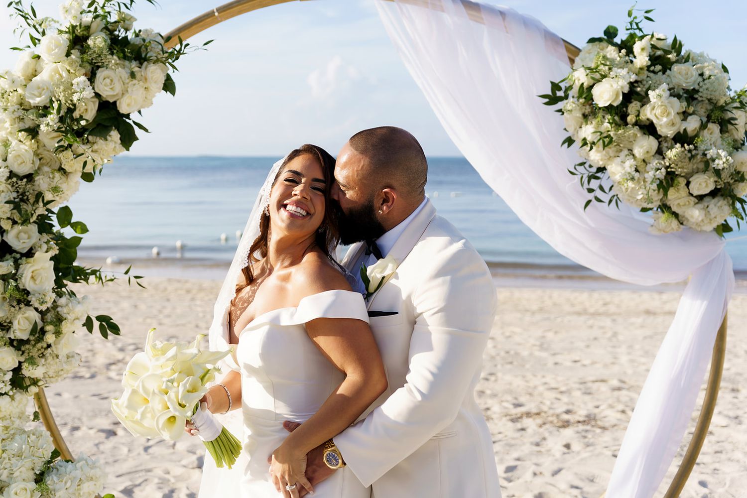 Couple embracing under floral arch on beach wedding, bride in white gown holding bouquet, groom in white suit.