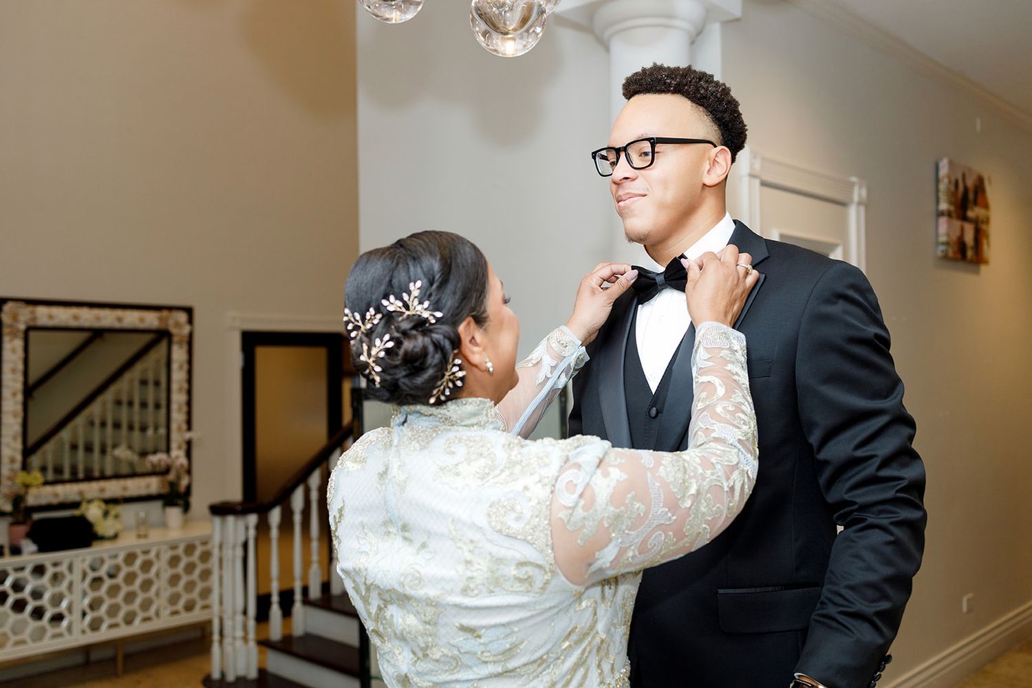 Mother adjusting groom's bow tie before wedding ceremony in elegant home setting.