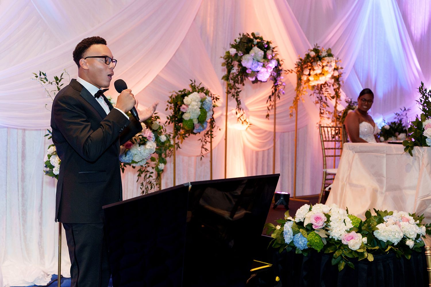 Elegant wedding reception emcee in black suit speaking into microphone beside floral arrangements and draped white backdrop.