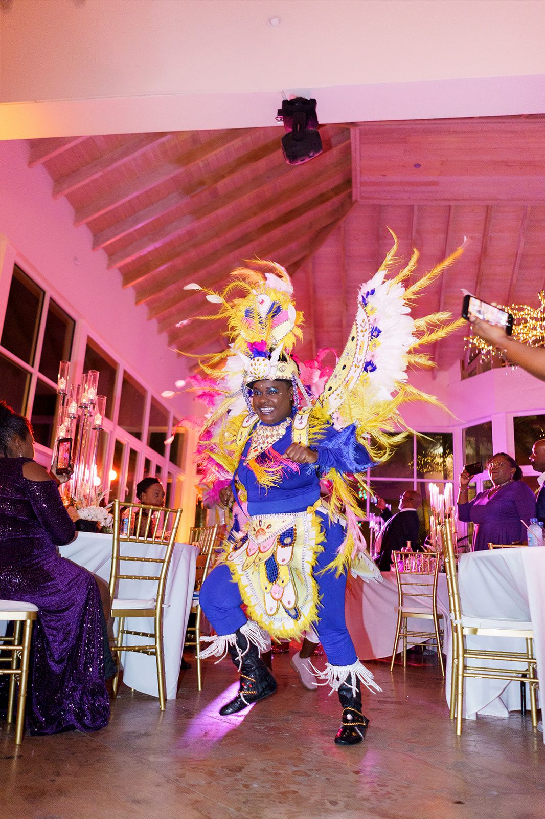 Colorful carnival dancer in feathered costume performing at elegant pink-lit event venue.