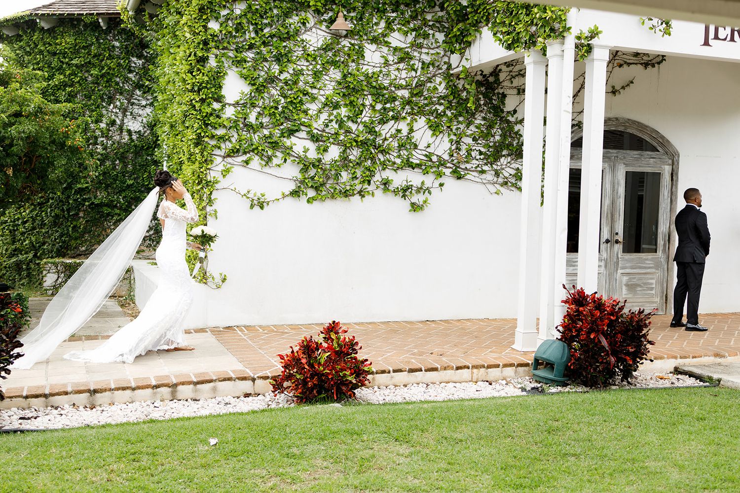 Bride in white gown with long veil stands outside elegant white villa as groom waits nearby.