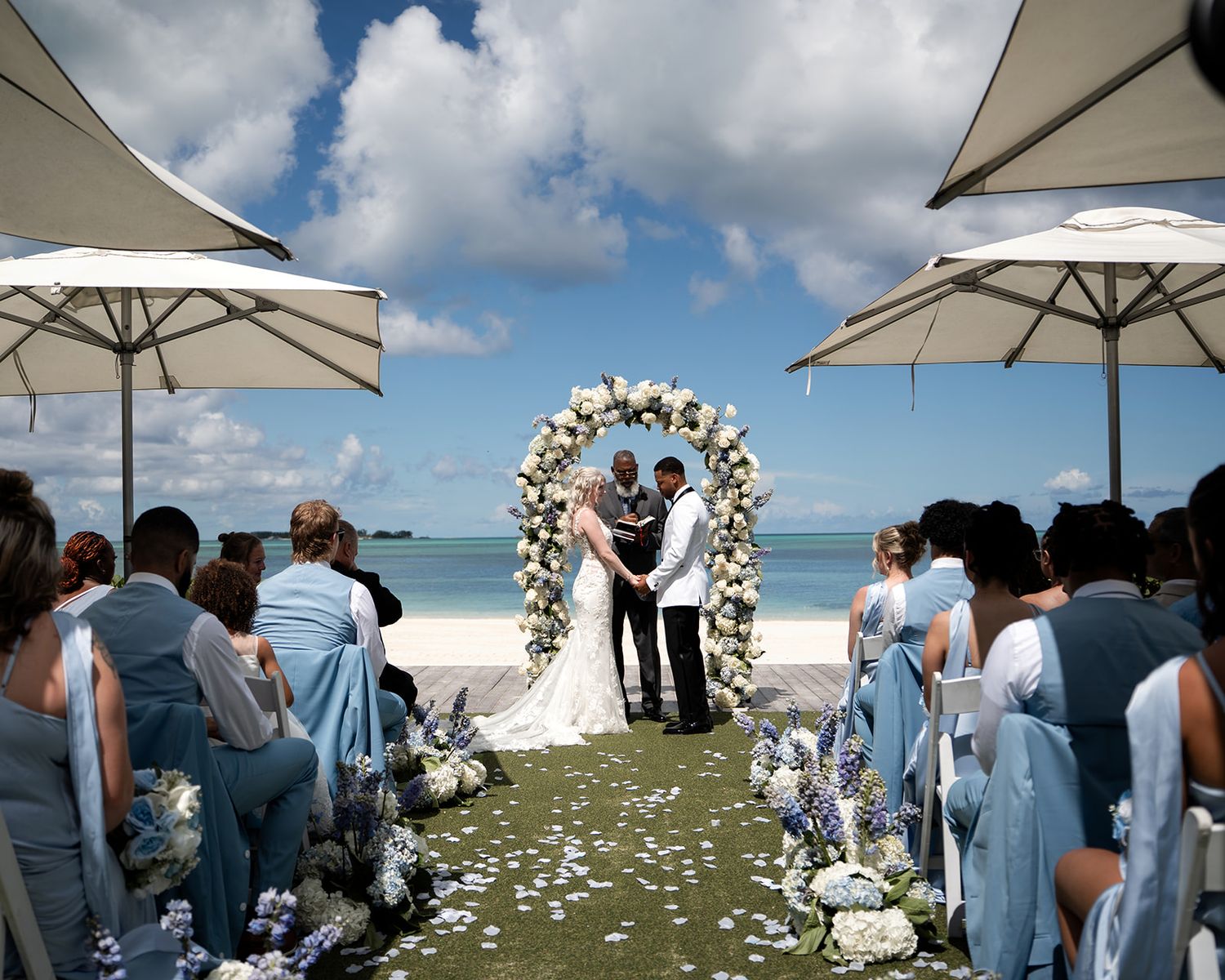 Romantic beach wedding ceremony with couple at floral arch, blue-dressed bridesmaids, white umbrellas, and ocean backdrop.