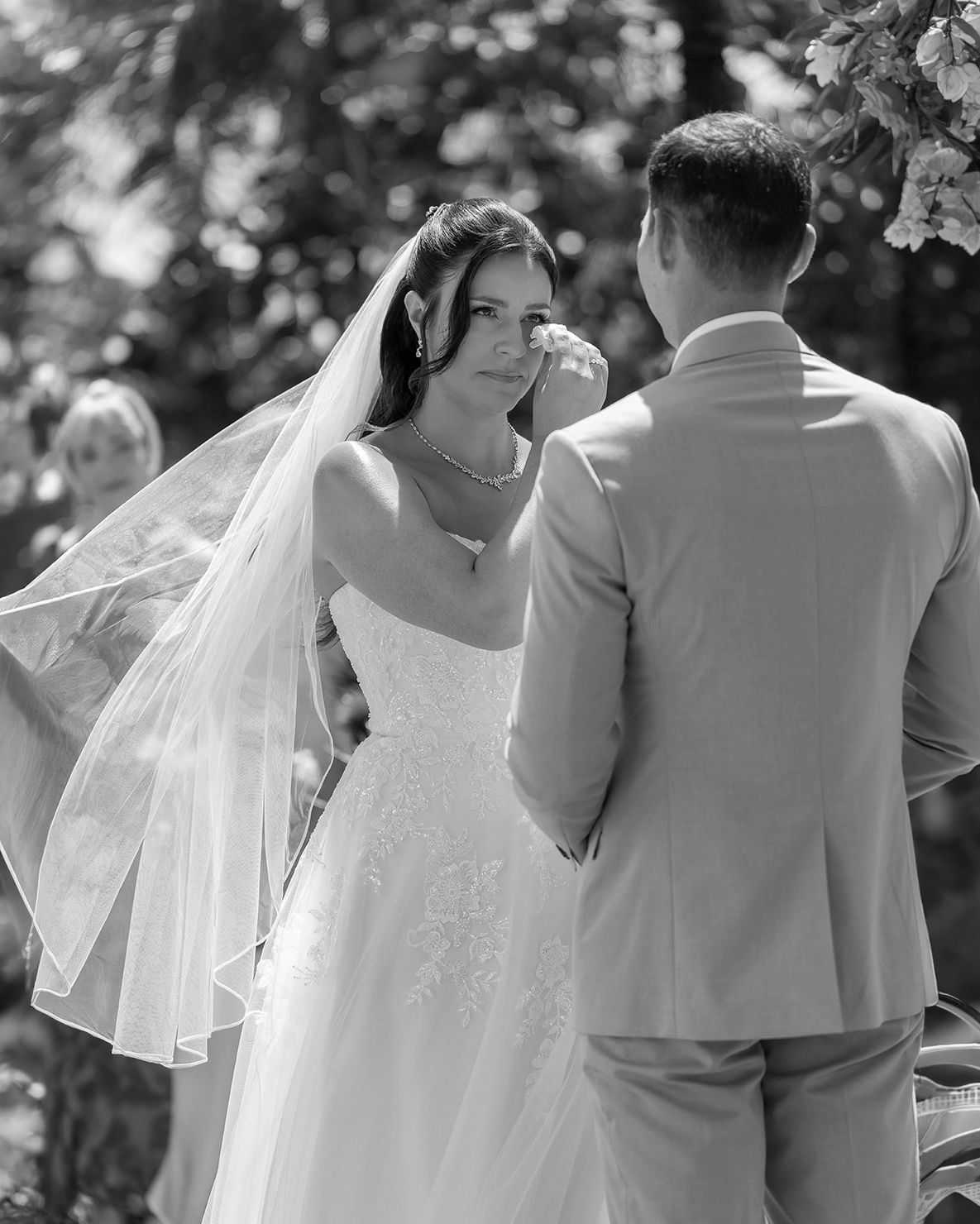 Black and white photo of a bride in a veil exchanging vows with a groom in a light suit at an outdoor wedding ceremony.