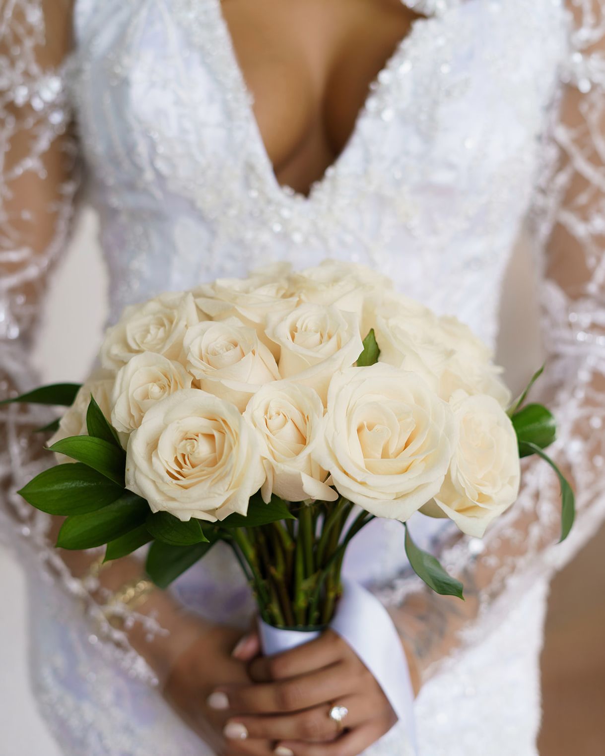Bride in beaded lace wedding gown holding elegant white rose bouquet with green leaves.