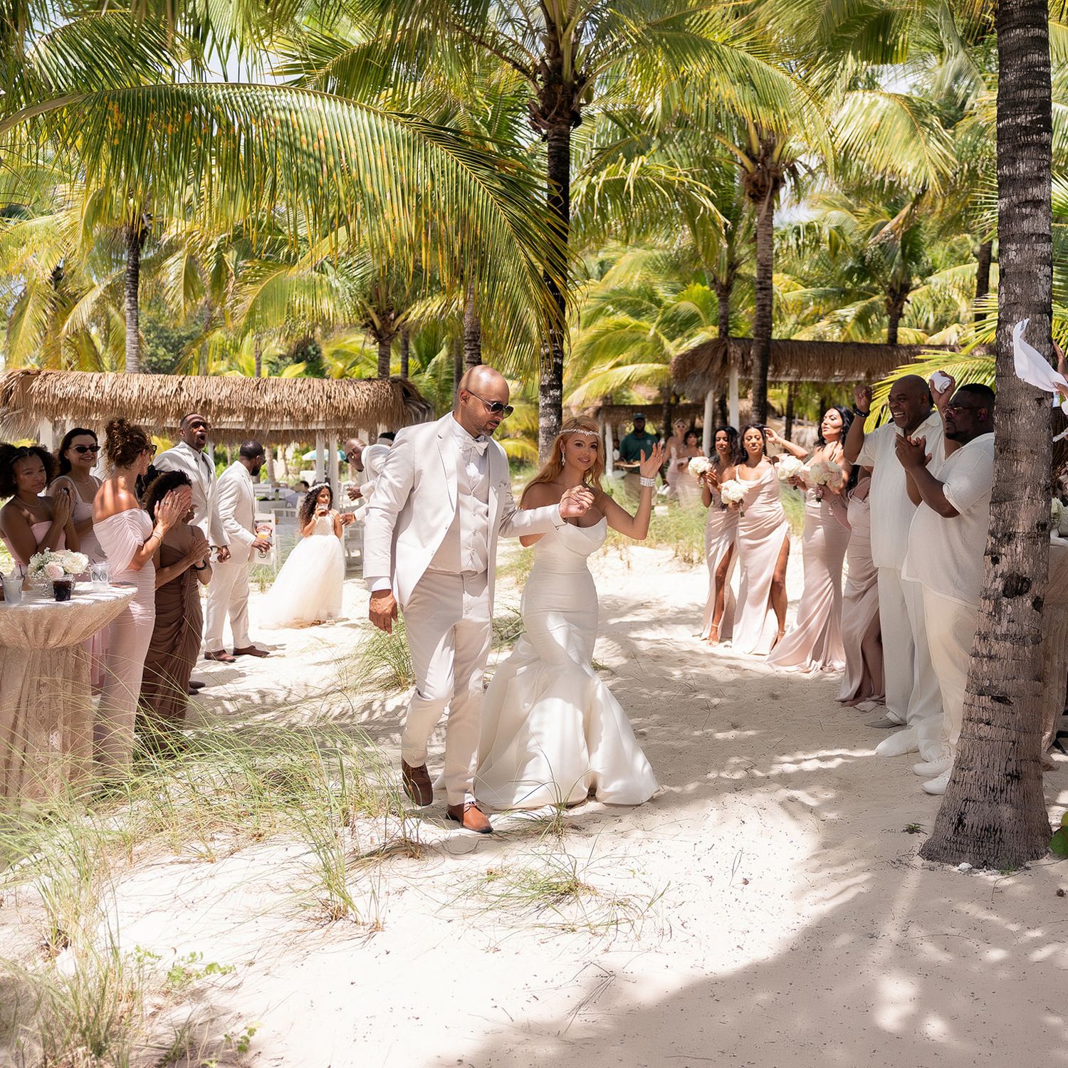 Bride and groom walk through sandy aisle lined with guests under tropical palm trees at outdoor beach wedding.