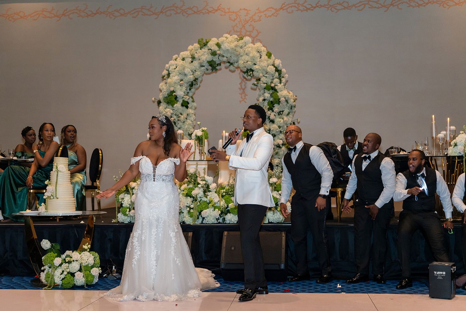 Groom serenades bride at elegant wedding reception with floral arch backdrop and guests in formal attire.