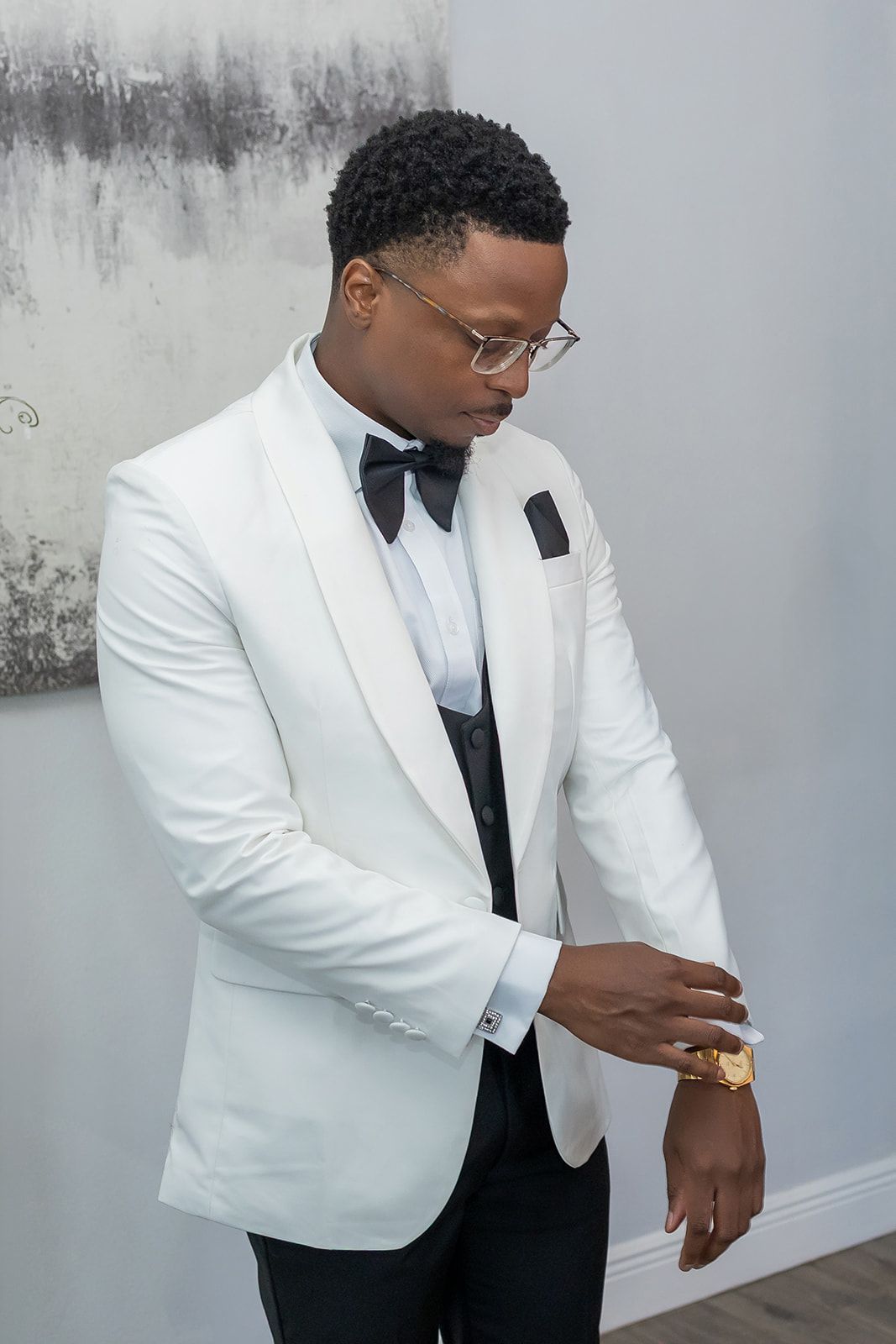 Elegant man in white tuxedo jacket with black bow tie and glasses, adjusting cufflinks at a formal event.