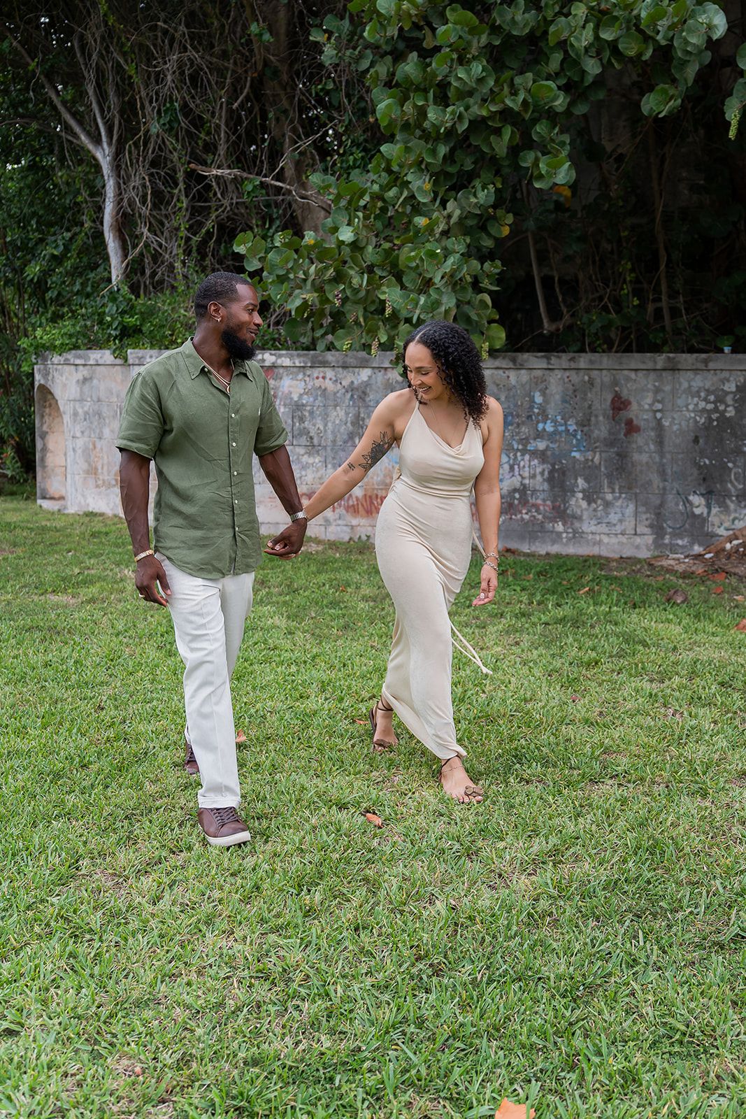 A couple walks hand-in-hand on green grass, smiling at each other, man in green shirt, woman in beige dress.
