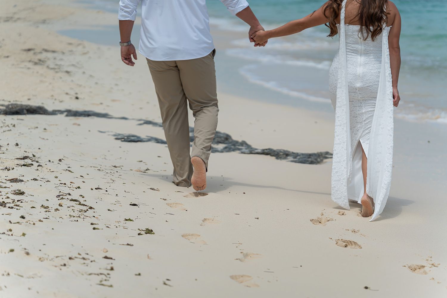 Couple holding hands walking barefoot on a white sandy beach, woman in white dress, man in casual attire.