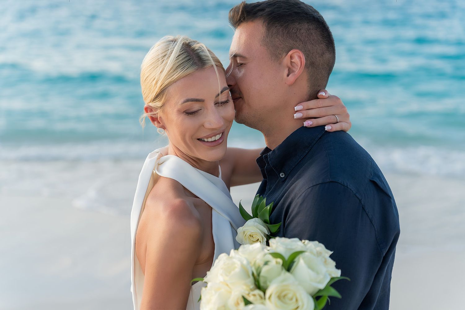 Couple sharing a romantic beach wedding moment, groom kissing bride's cheek as she holds white roses bouquet.