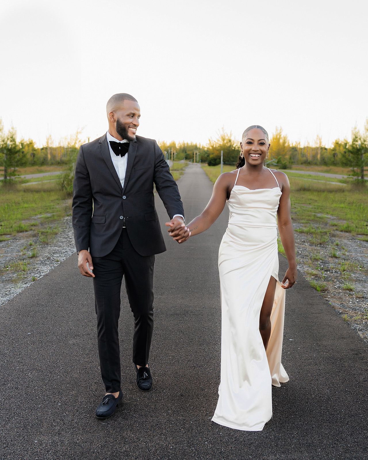 A couple in wedding attire holds hands walking down a road, bride in white gown and groom in black tuxedo.