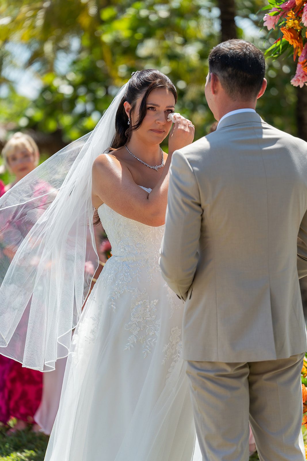 Bride in white gown and veil wiping tears while exchanging vows with groom at outdoor wedding.
