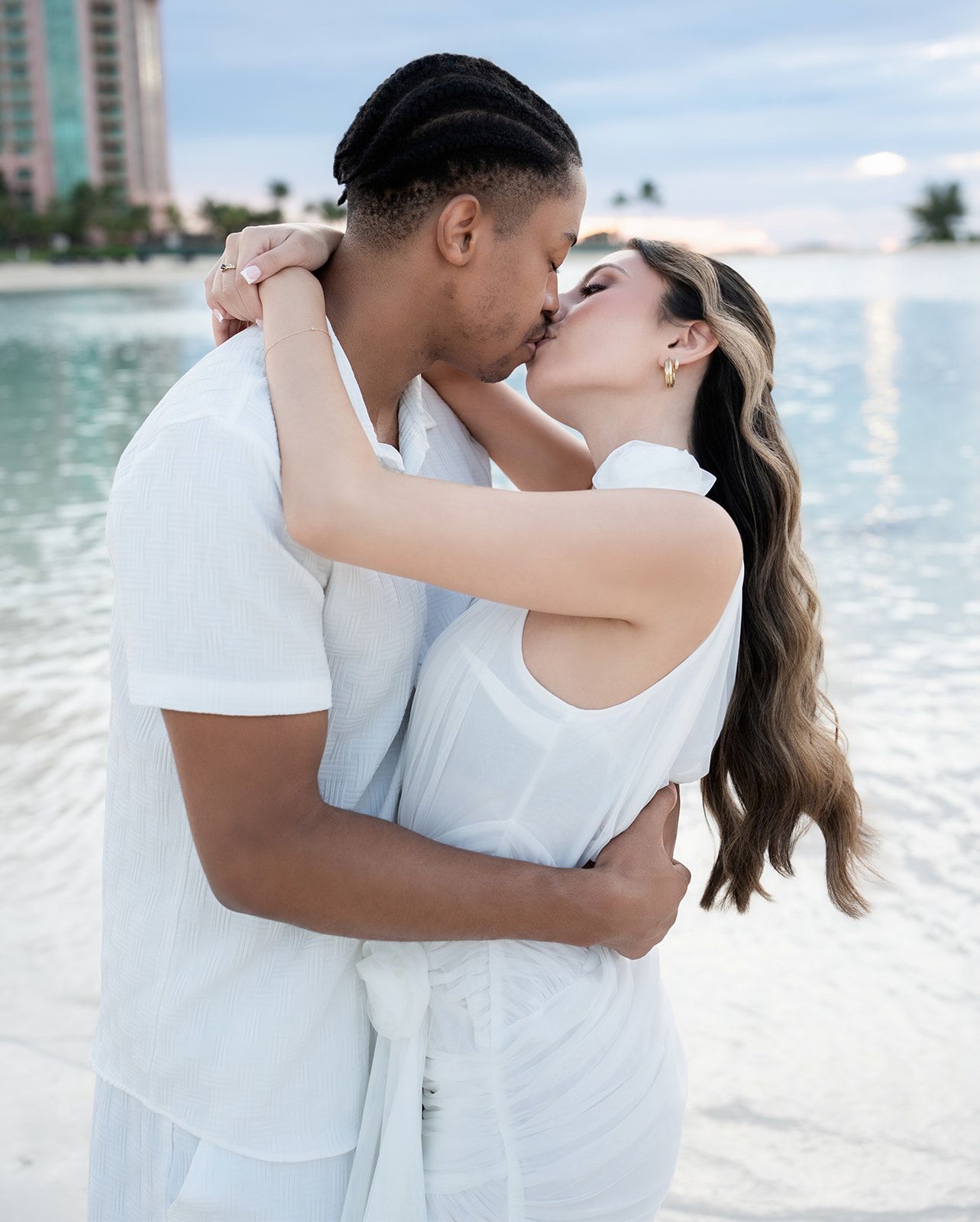 Couple in white outfits sharing a romantic kiss on a tropical beach with turquoise water and resort in background.