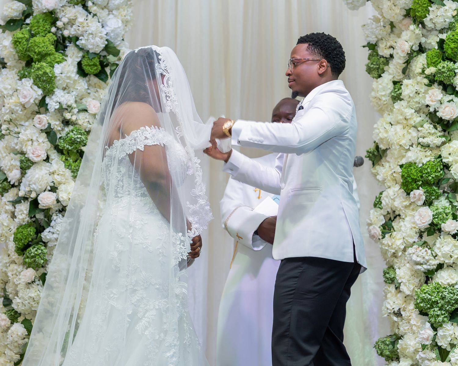 Groom in white tuxedo lifts bride's veil at elegant floral wedding altar adorned with white roses.