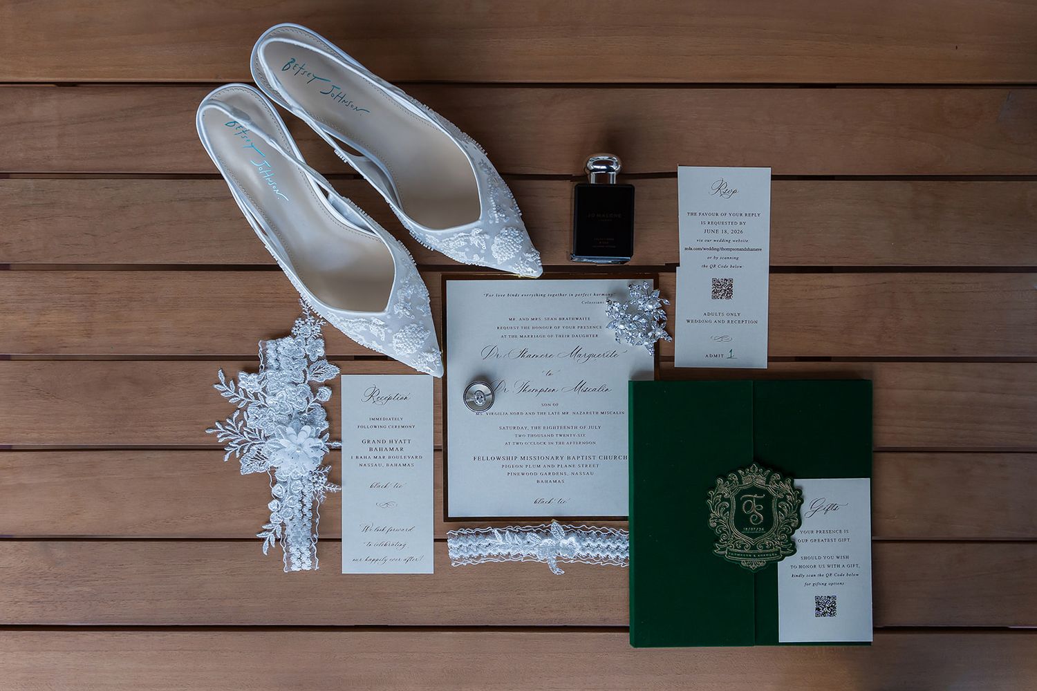 Elegant wedding flat lay featuring lace bridal shoes, garter, invitation suite, rings, and green monogrammed book on wood.