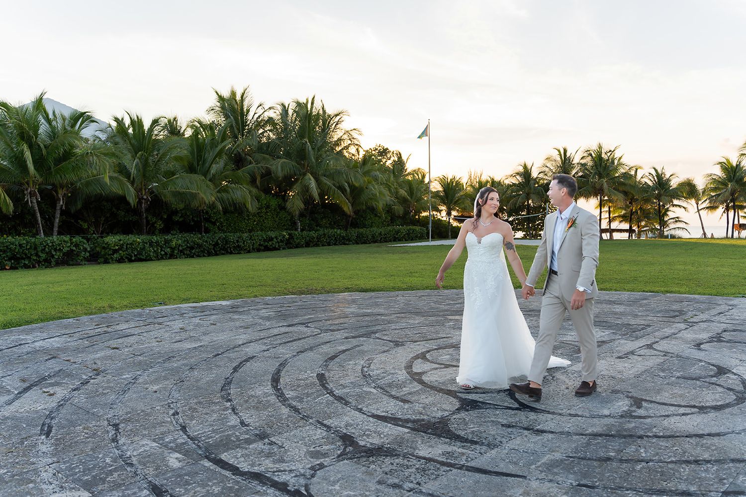 Bride and groom walk hand-in-hand on a stone labyrinth surrounded by tropical palm trees at a destination wedding.