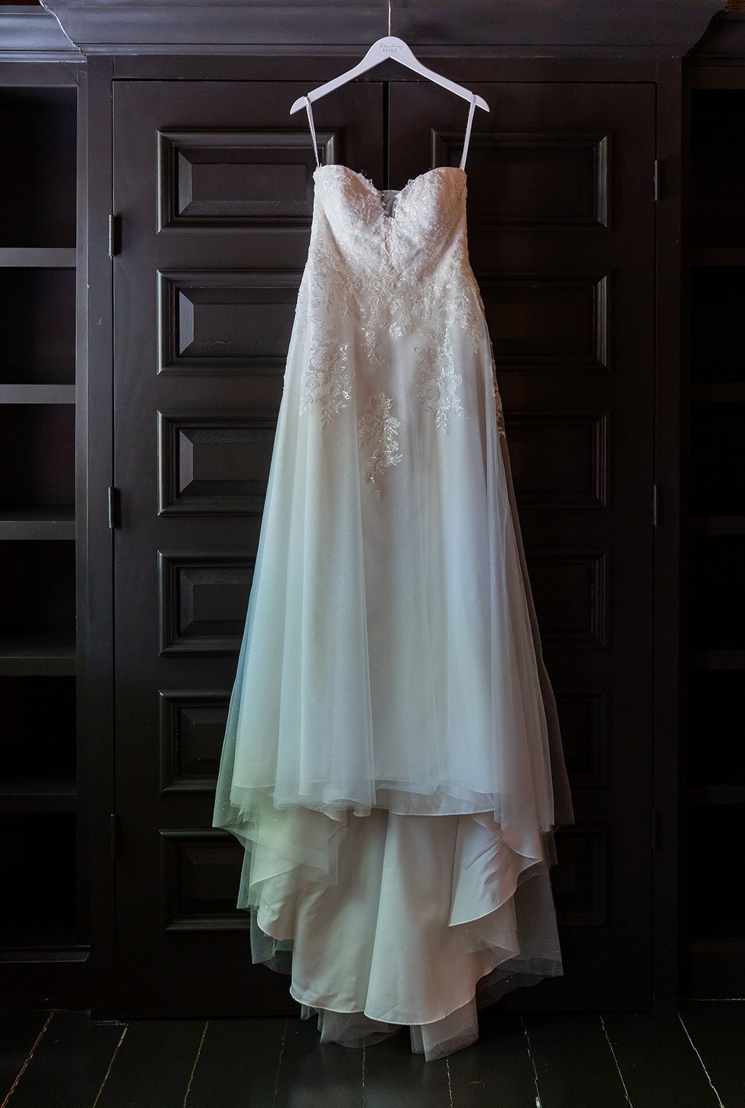 Elegant strapless lace bodice wedding gown with flowing chiffon skirt hanging on a hanger against dark wood doors.