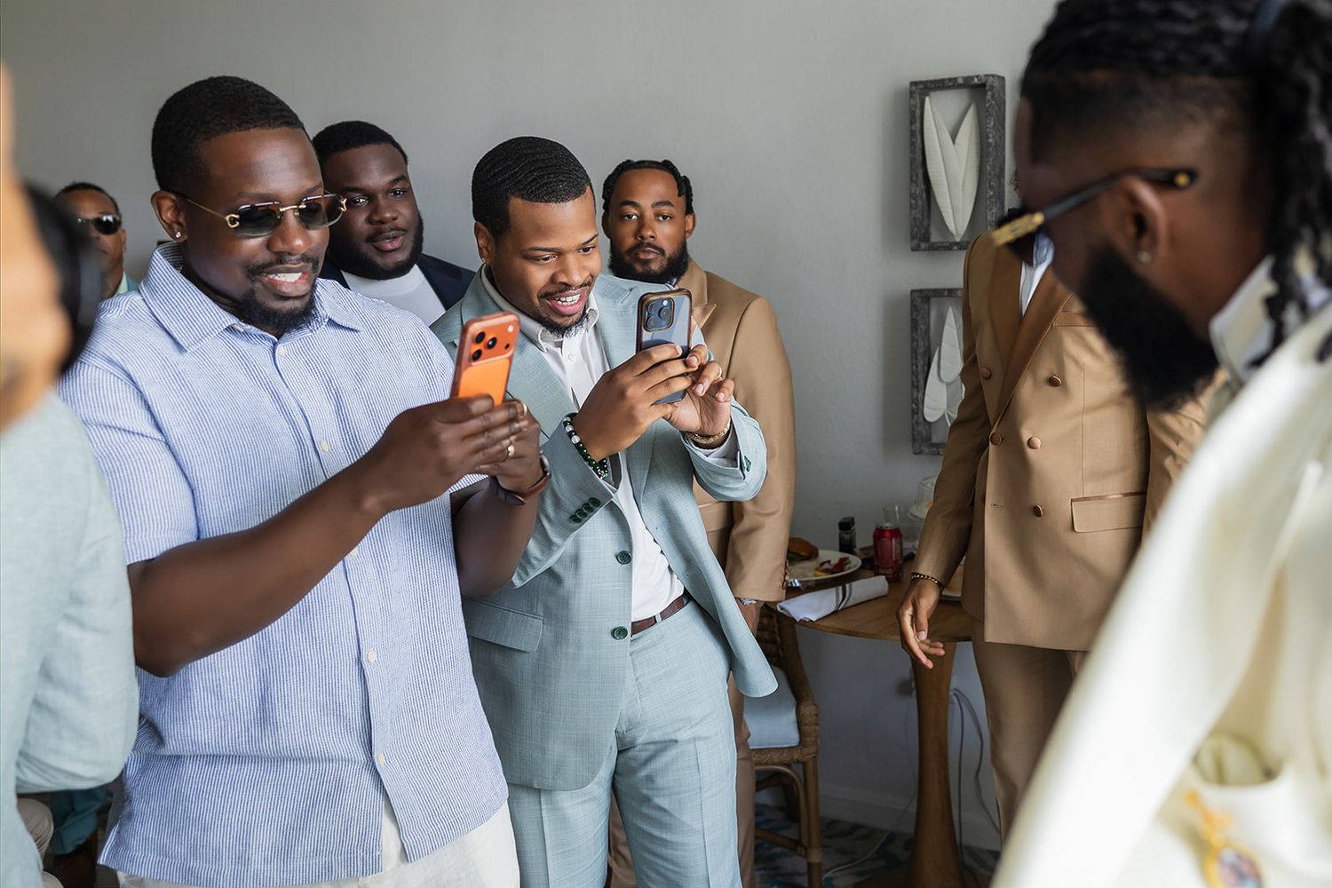 Group of well-dressed men at a wedding celebration, smiling and taking photos on smartphones indoors.