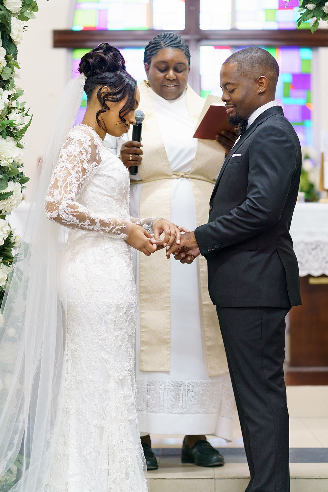 Bride and groom exchange rings at church altar during wedding ceremony, officiated by female minister holding microphone.
