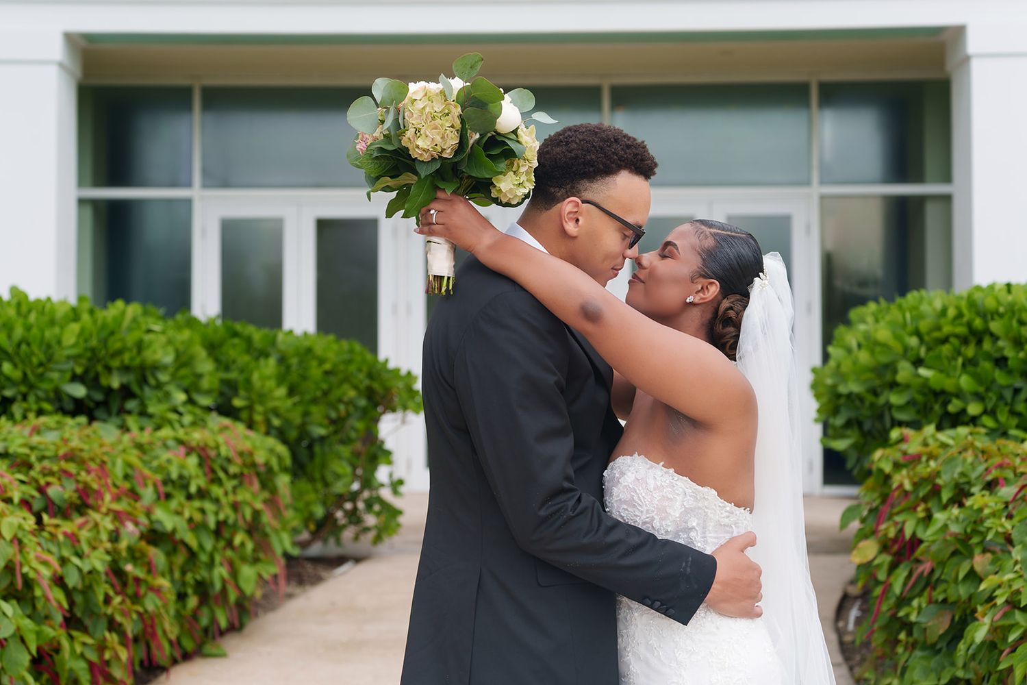 A newlywed couple shares a romantic kiss outdoors, the bride in a white gown holding a bouquet while the groom wears a black suit.