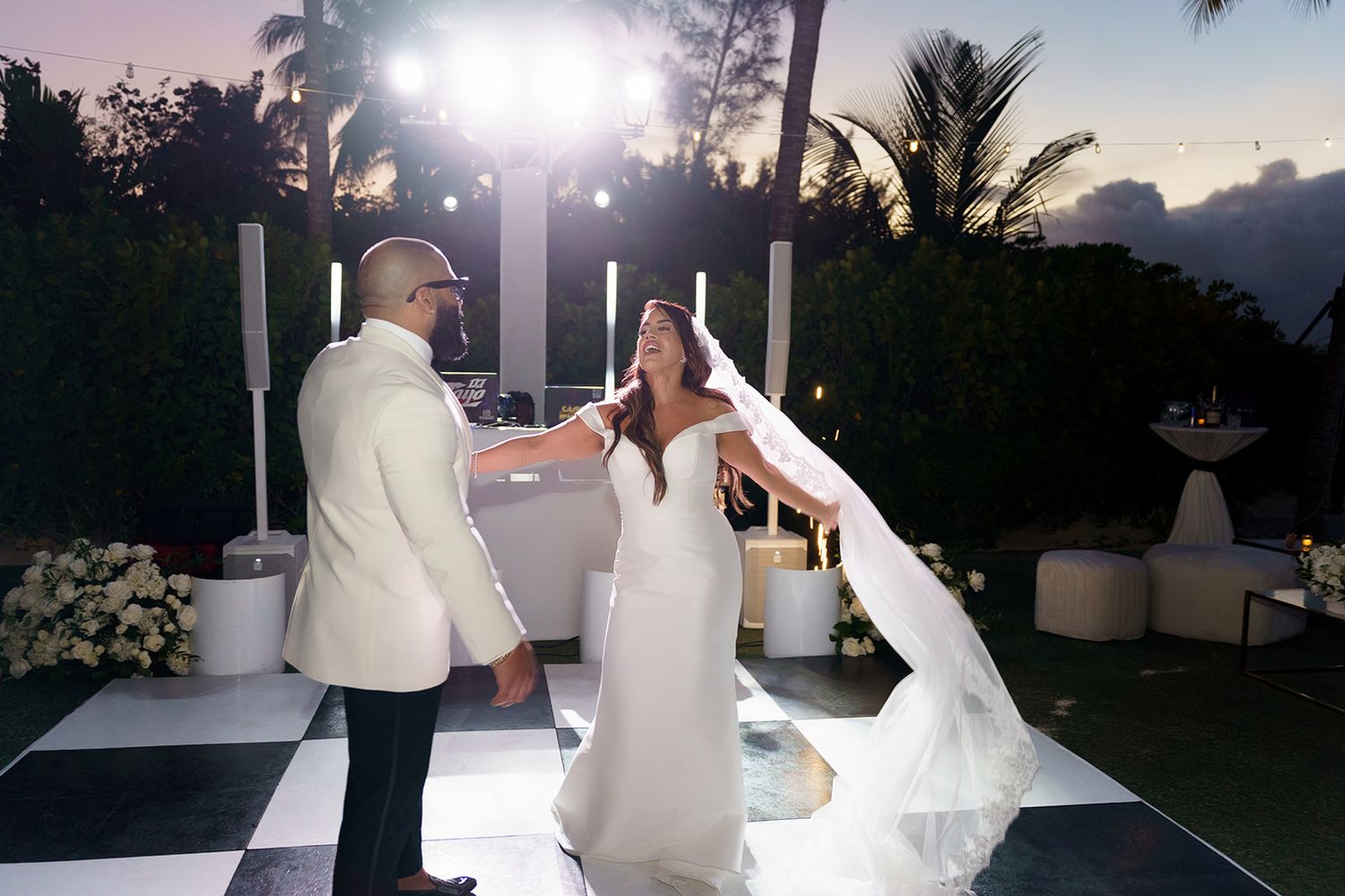 Bride and groom share first dance on checkered floor at elegant outdoor evening wedding reception with palm trees.