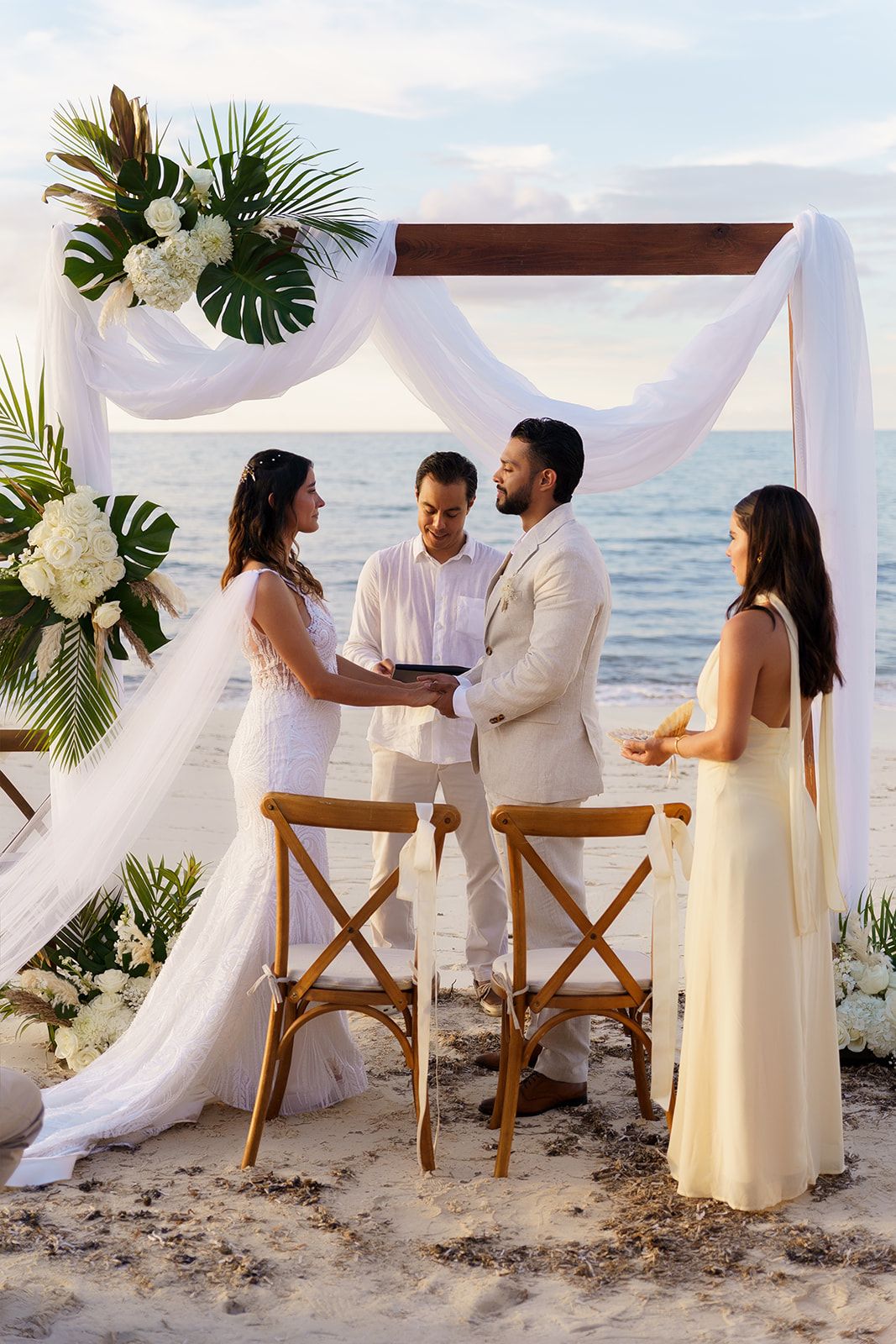 Couple exchanges vows at elegant beach wedding ceremony under white draped arch with tropical floral arrangements at sunset.