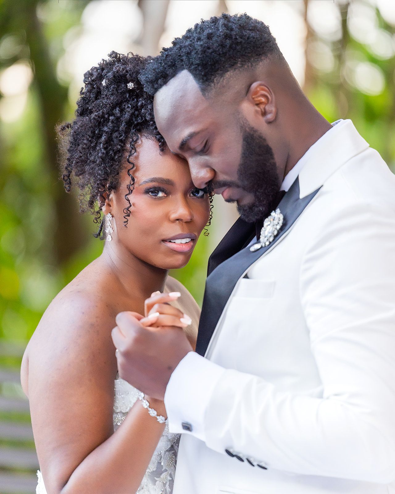 A bride and groom in elegant wedding attire share an intimate moment, touching foreheads tenderly outdoors.