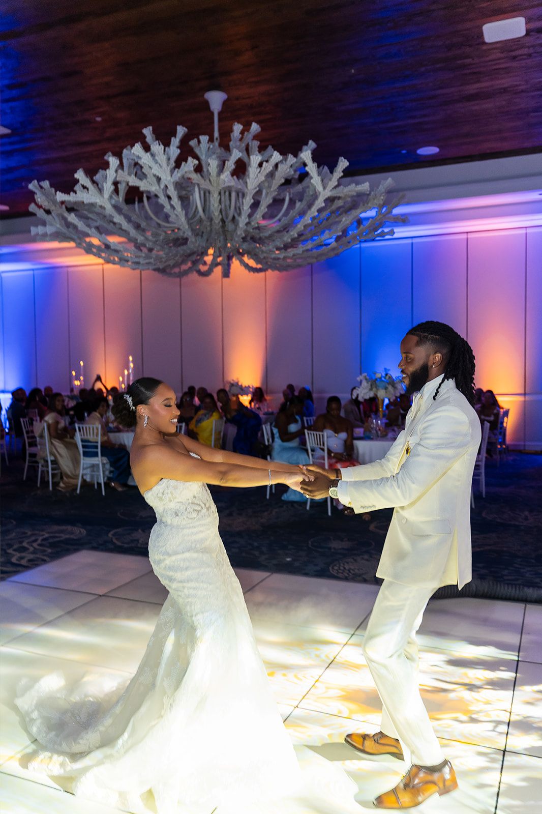 Bride and groom share first dance under elegant chandelier in decorated ballroom with blue uplighting.