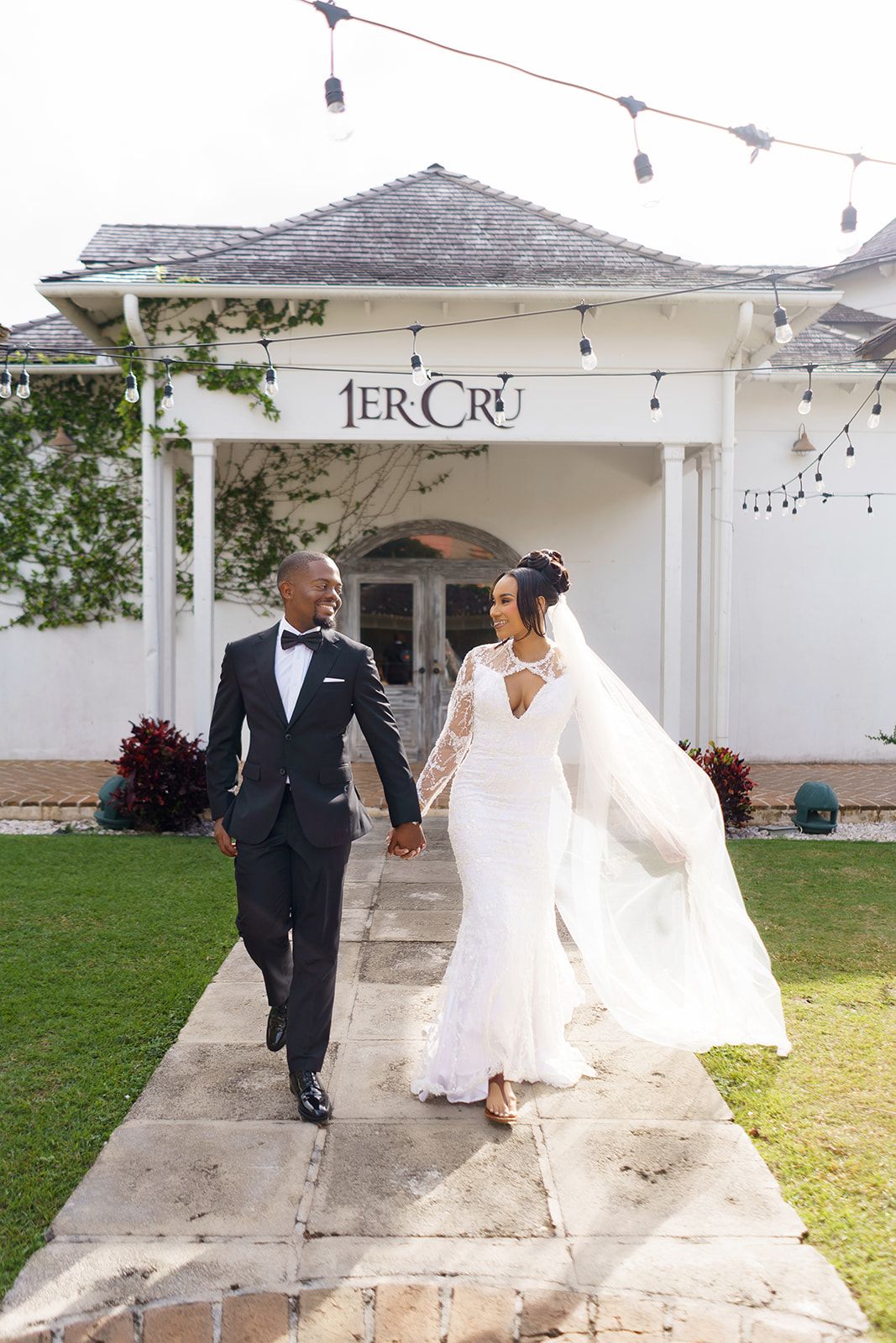 Bride and groom holding hands walking in front of 1er Cru venue, bride in lace gown with veil flowing.
