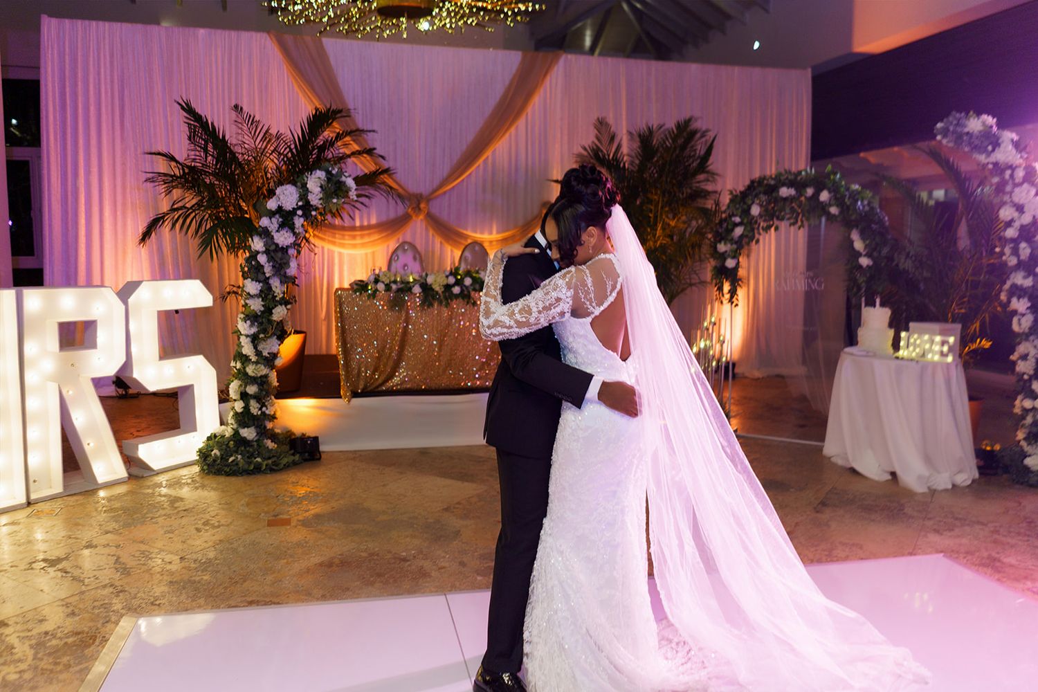 A bride and groom share their first dance at an elegant wedding reception with purple lighting.