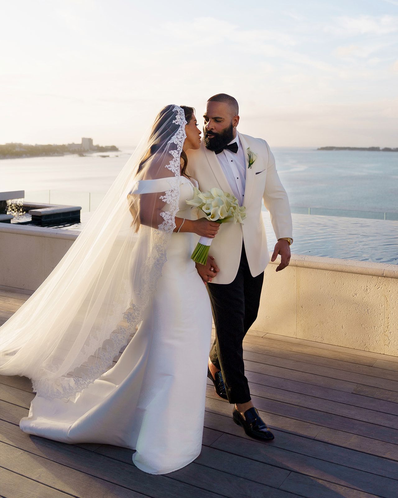 Bride and groom share a romantic moment on a waterfront terrace, bride in white gown with veil, groom in white tuxedo jacket.