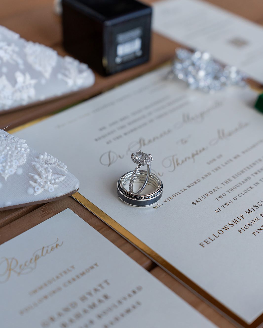 Elegant wedding stationery suite with gold-trimmed invitations, diamond engagement ring, and wedding bands on a wooden surface.